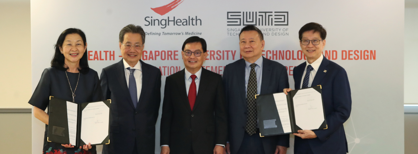 Five people in suits holding documents, standing in front of a SingHealth and SUTD banner.